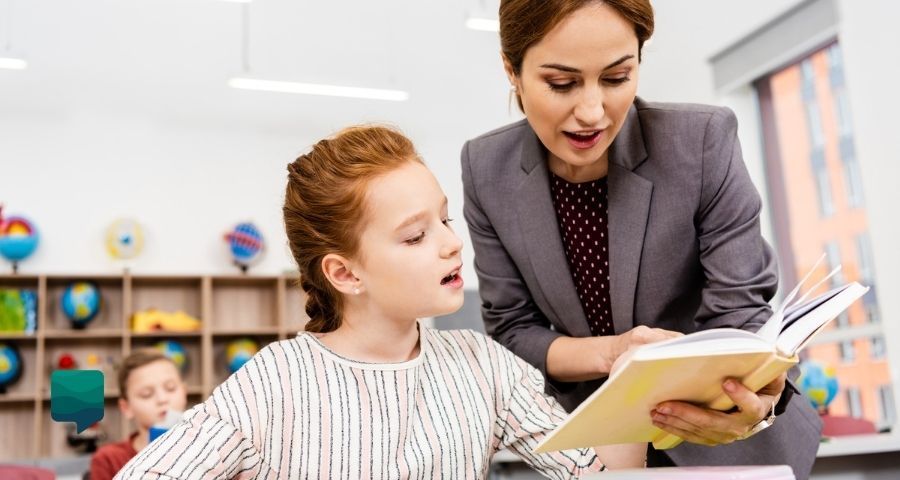 Como preparar aula de inglês para iniciantes: foto de uma professora apontando para o livro enquanto aluna l