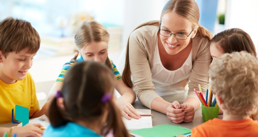 Professora na mesa em meio aos alunos ouvindo-os e sorrindo dando uma aula de inglês inesquecível.