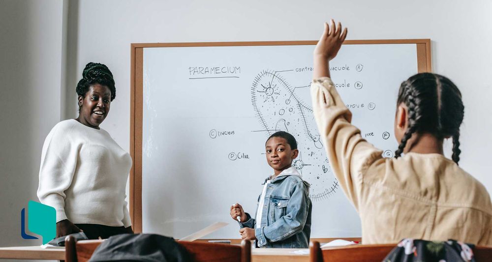 Professora e aluno em frente a um quadro branco, enquanto uma aluna, sentada, levanta a mão