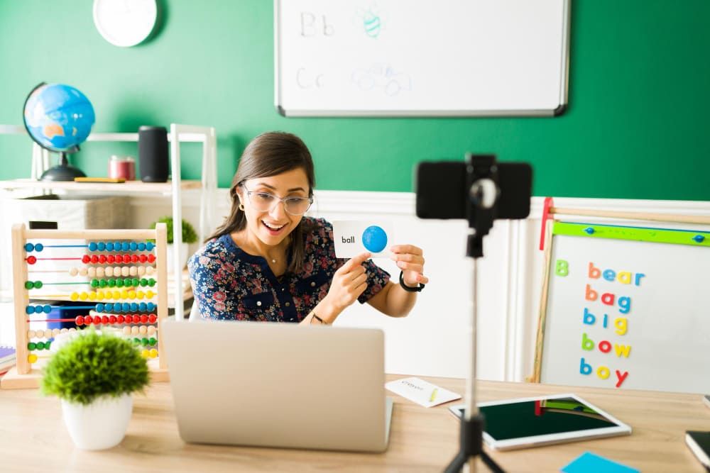 Professora em frente a um computador e uma câmera gravando uma vídeoaula para o seu curso online de inglês