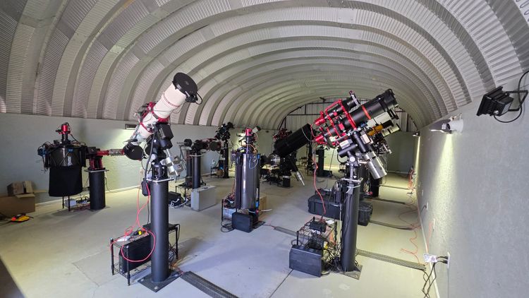 A white German equatorial mount carrying a refractor on a workbench, cabled and ready for a remote observatory.
