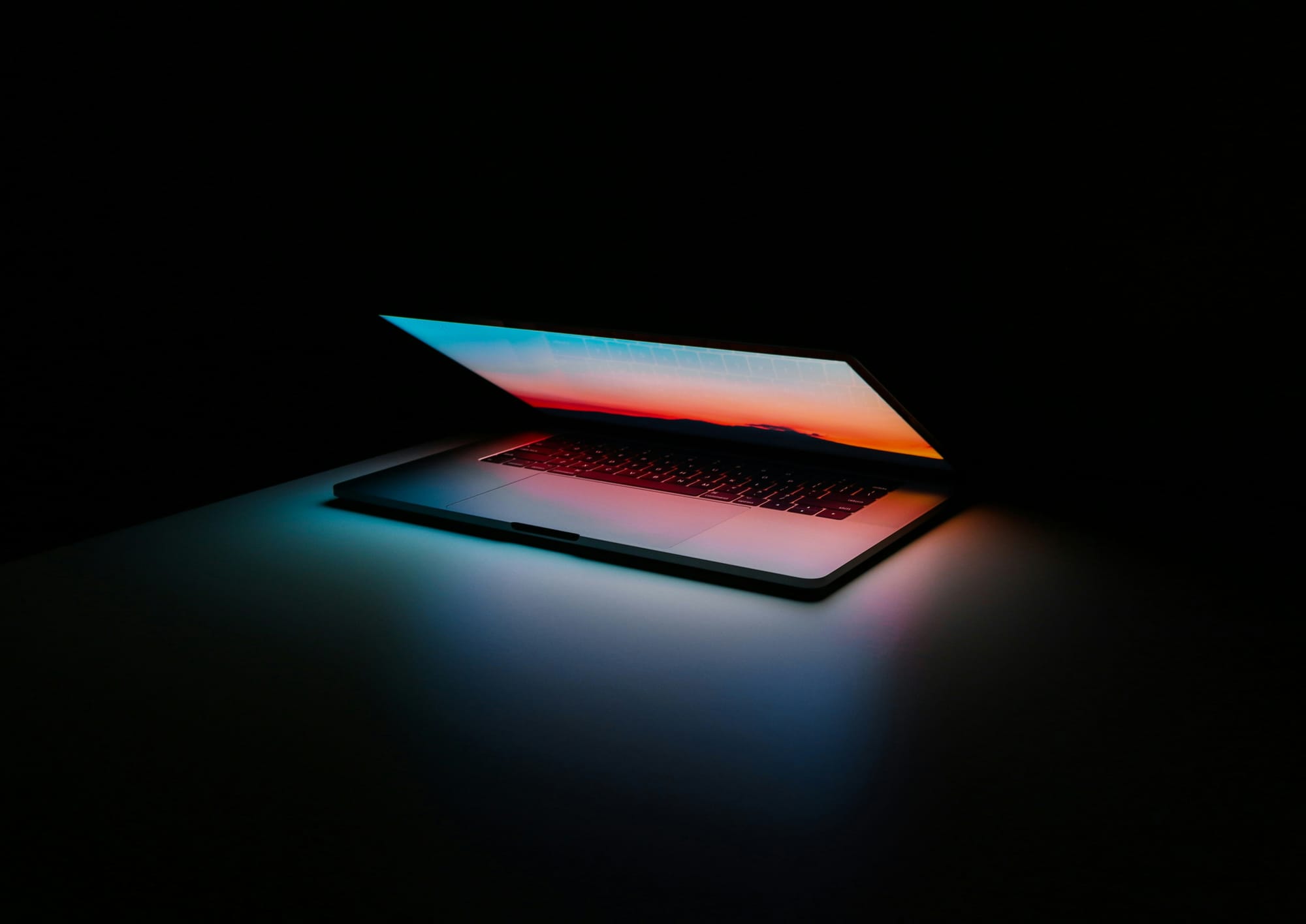 A silver, apple brand laptop sits on a dark surface in a darkened space. The computer is partially opened, with a soft multi-colored background showing blues and pinks.