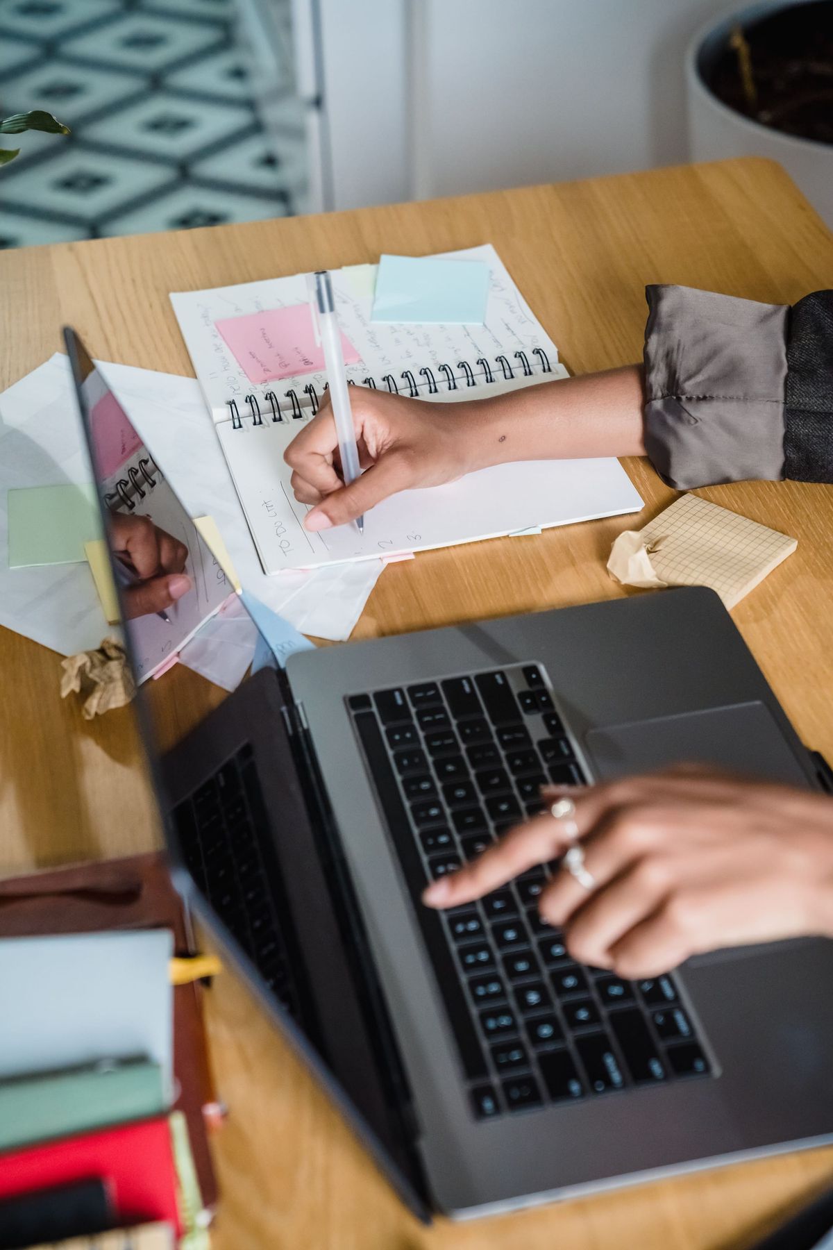 A laptop sits on a desk with notes around it. A woman jots down ideas, while pointing at the computer screen.