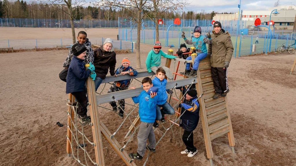 Kasvattajat auttavat toiminnassa mukana olevia lapsia koulussa, kotona ja vapaa-ajalla 12 vuoden ajan. Kuvassa kasvattaja ja joukko lapsia leikkimässä kiipeilytelineessä.
