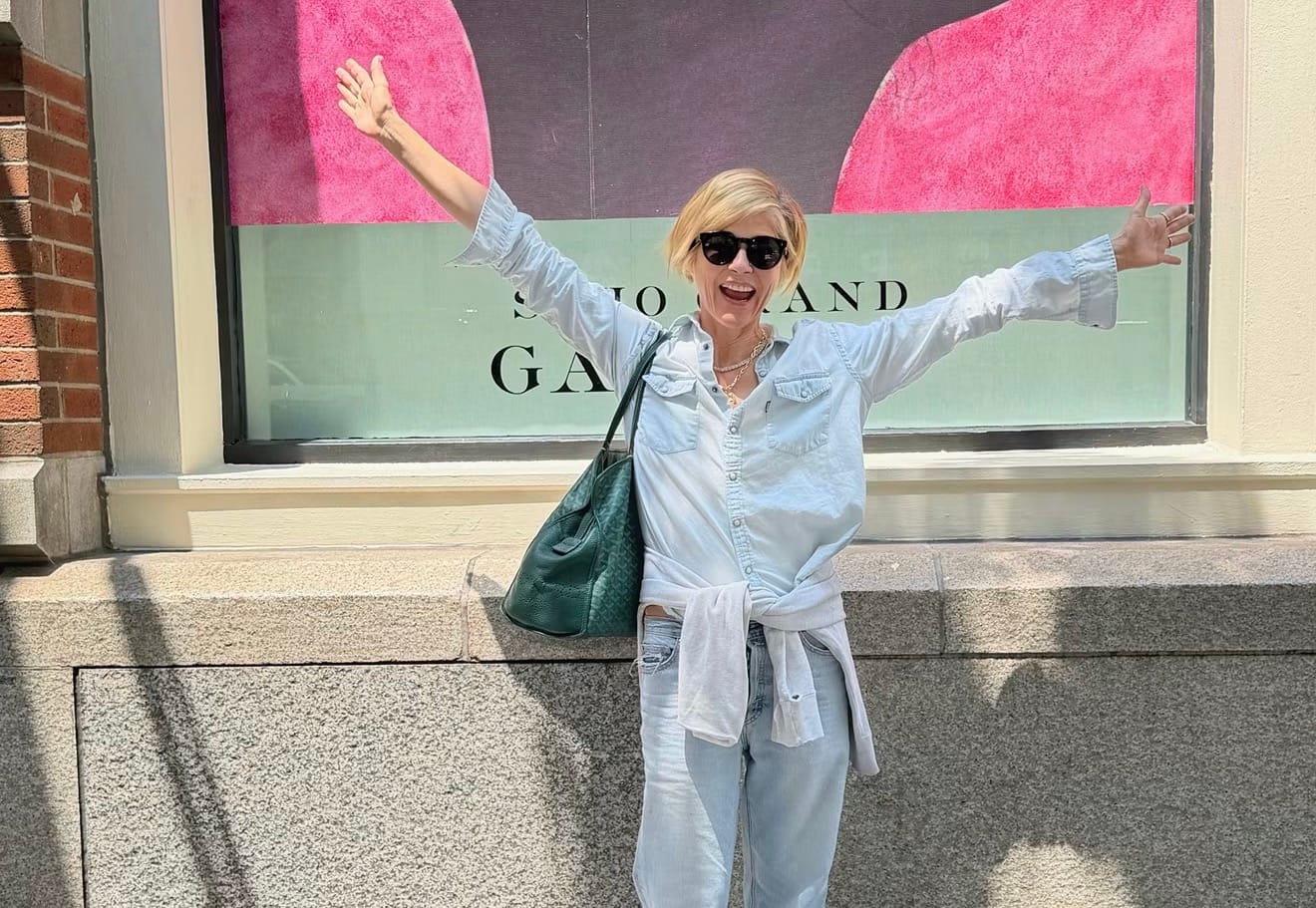 Woman in sunglasses and a denim shirt smiling with her arms outstretched on a city sidewalk