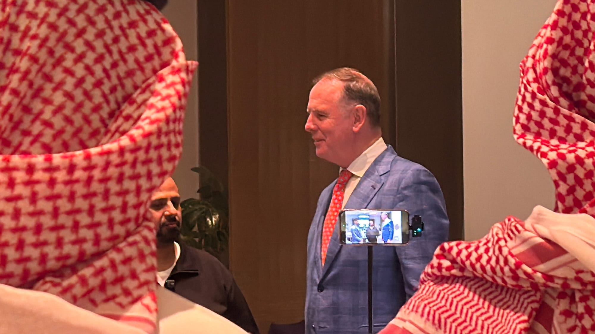 Riyadh Air CEO Tony Douglas in a blue suit, being filmed on a phone, framed by red-and-white headscarves in the foreground