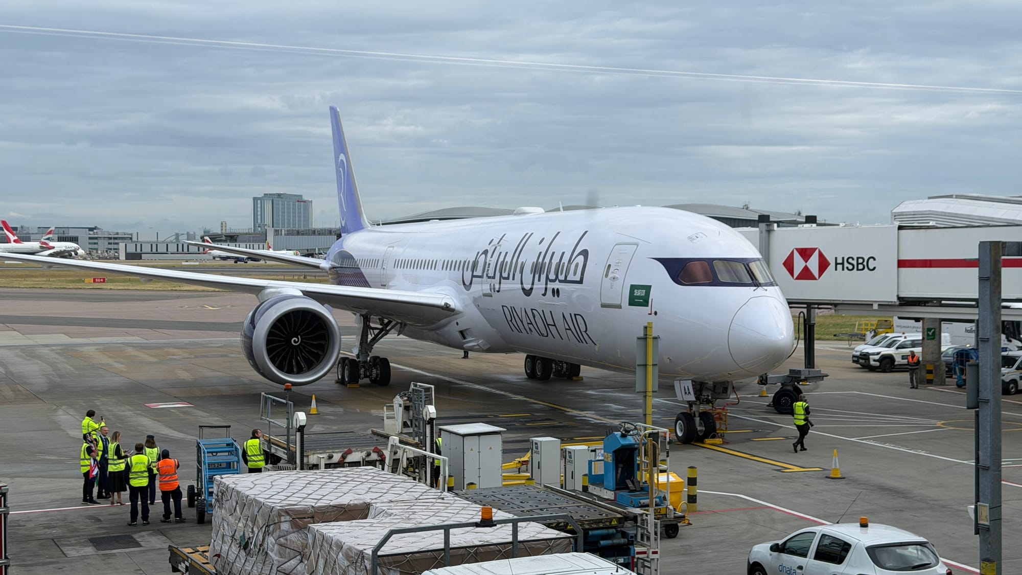 Riyadh Air Boeing 787-9 parked at a London Heathrow gate with ground crew unloading cargo