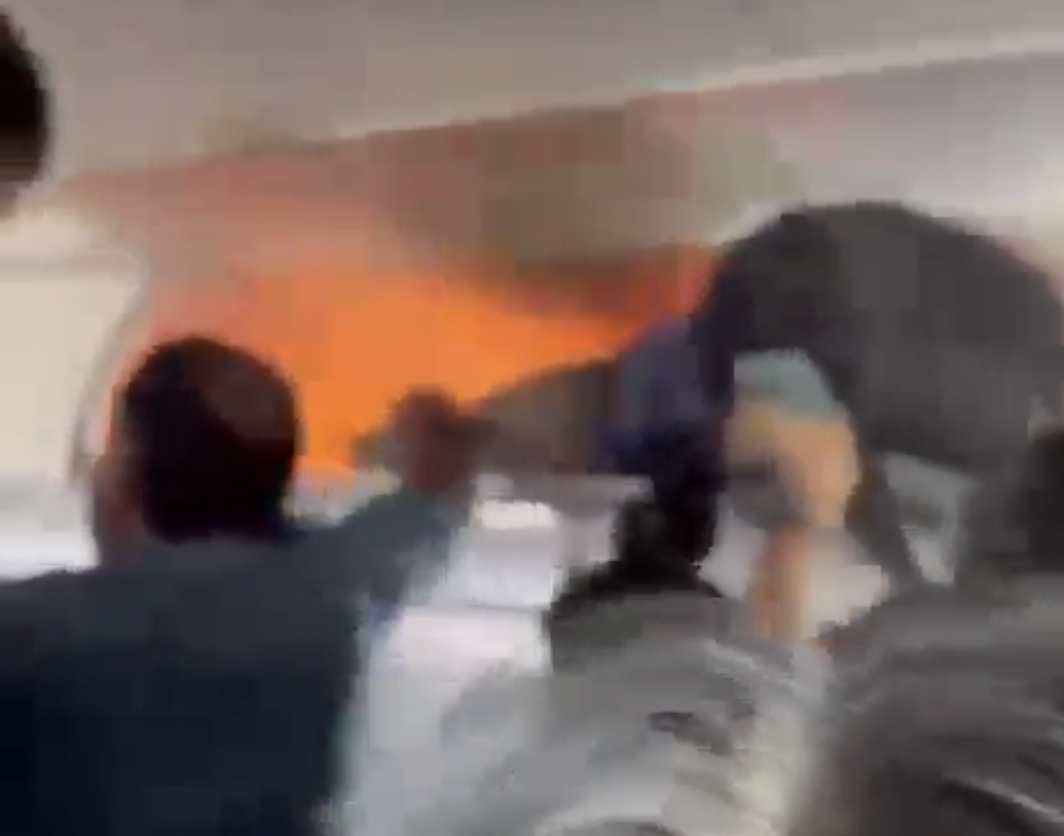 Blurry video still of passengers reaching toward orange flames in an airplane overhead bin