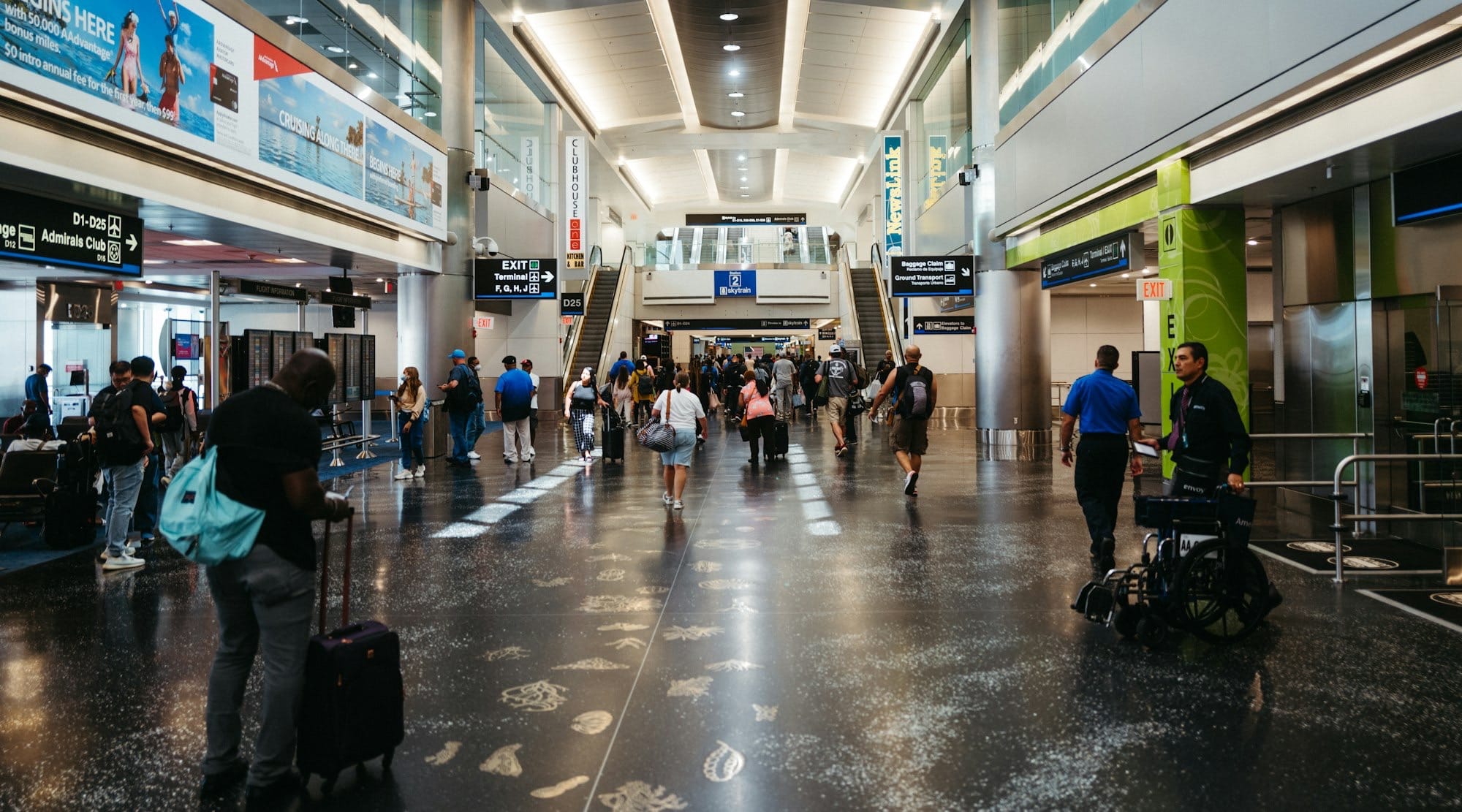 Busy concourse at Miami International Airport with travelers pulling suitcases past shops and an Admirals Club sign
