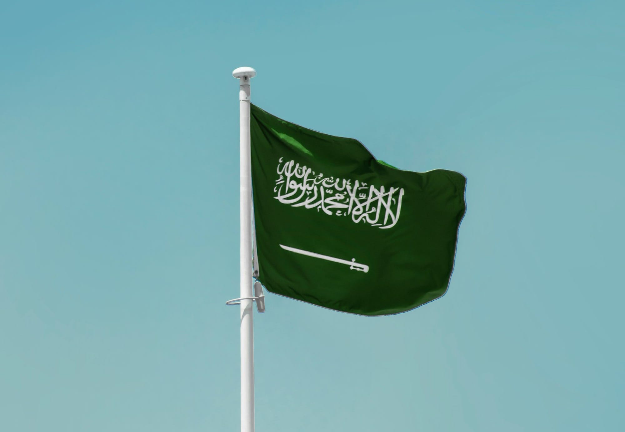 Green Saudi Arabian flag with white Arabic script and a sword, flying against a blue sky