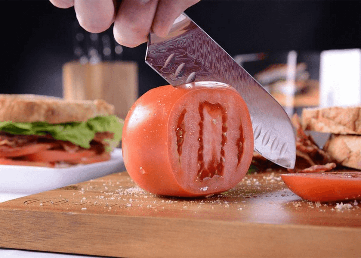 A Santoku knife slicing a tomato on a cutting board with a sandwich in the background.