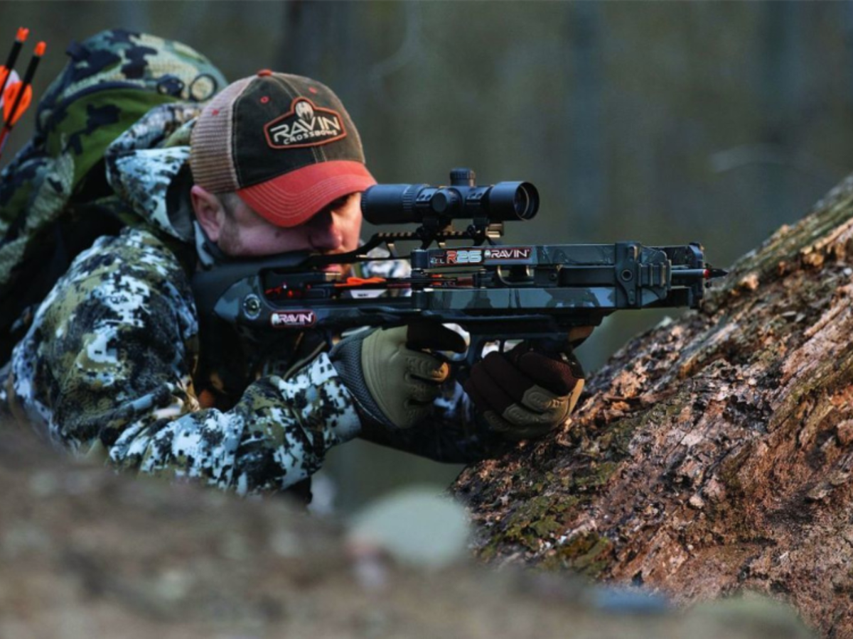 A man in winter camo with his Ravin cocked, and aimed at his target while in a kneeling position shooting off a tree.