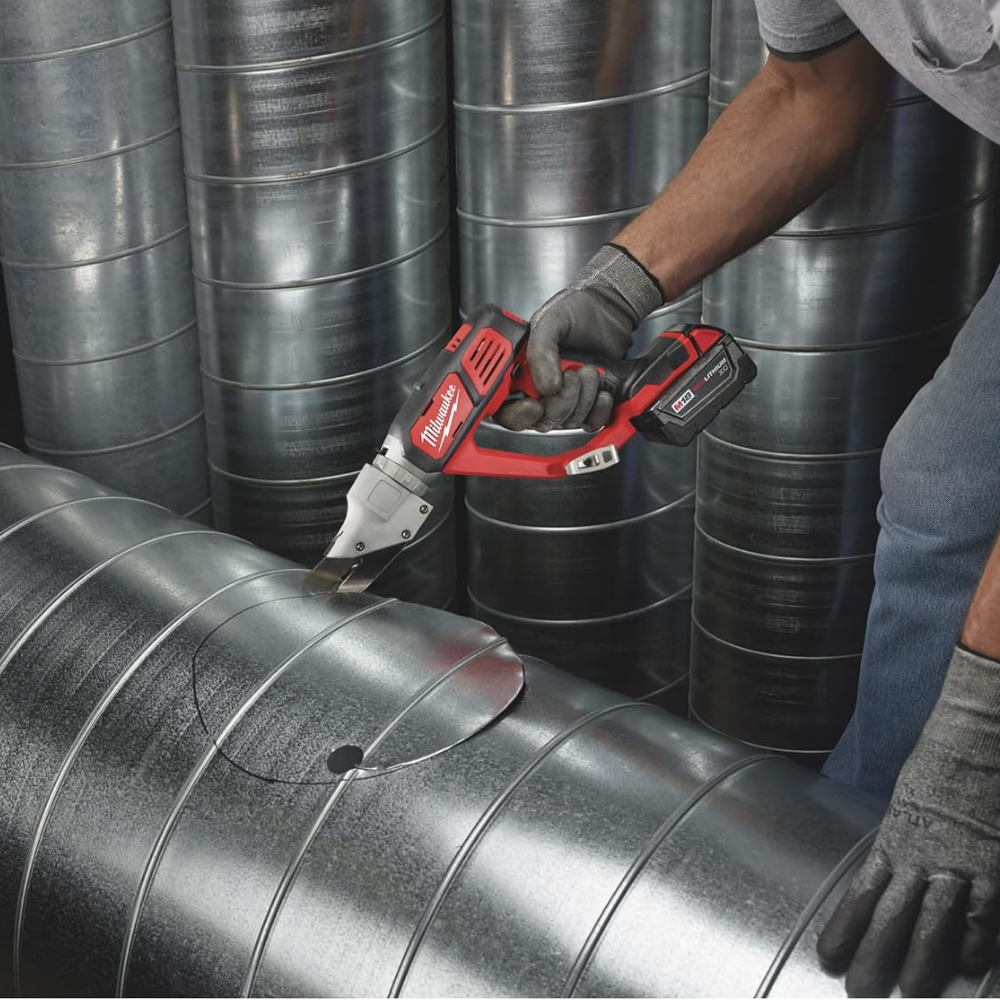 A man using M18 cordless shears to cut a circle in a large metal pipe.