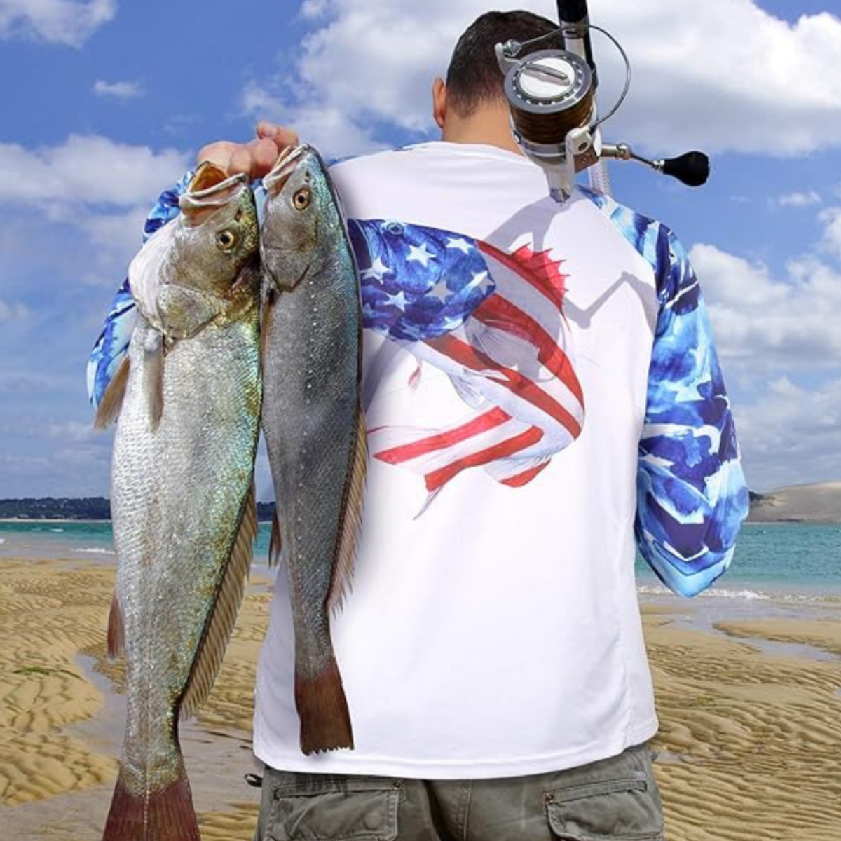 A man carrying 2 fish and a fishing pole walking on a beach wearing a fishing shirt with long sleeves