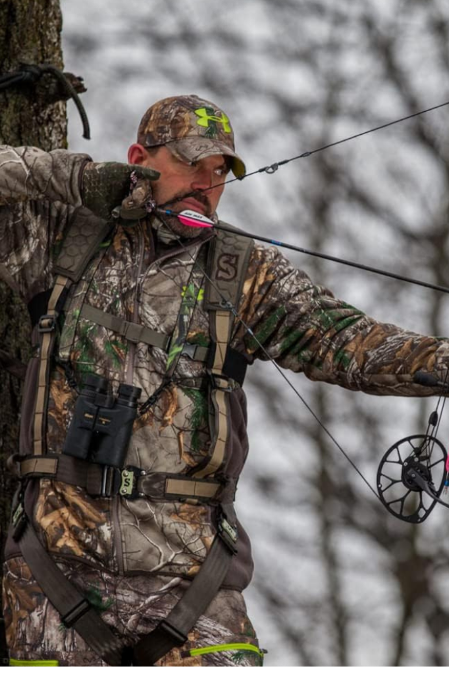 A man with a bow drawn from his tree stand using a vest style safety harness