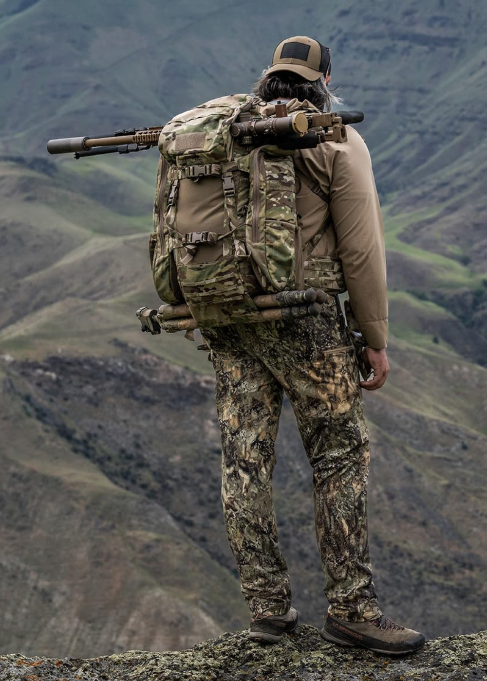 A man with a tactical backpack in camo looking off from the crest of a ridge of rocks