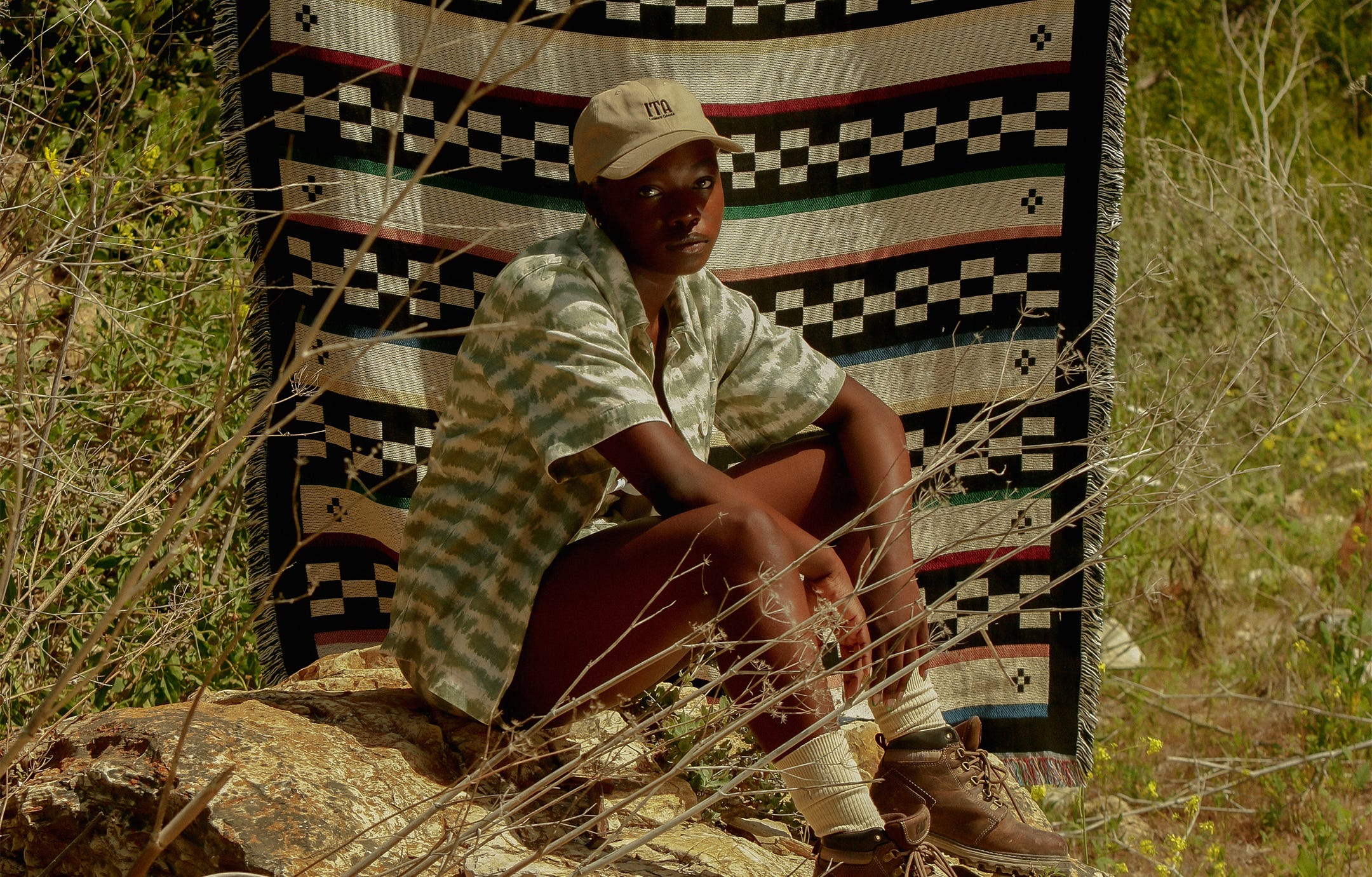 A female model wearing a striped shirt, hiking boots, and a tan ITA Lesiure ball cap sits on a rock in front of a patterned ITA Leisure outdoor blanket 