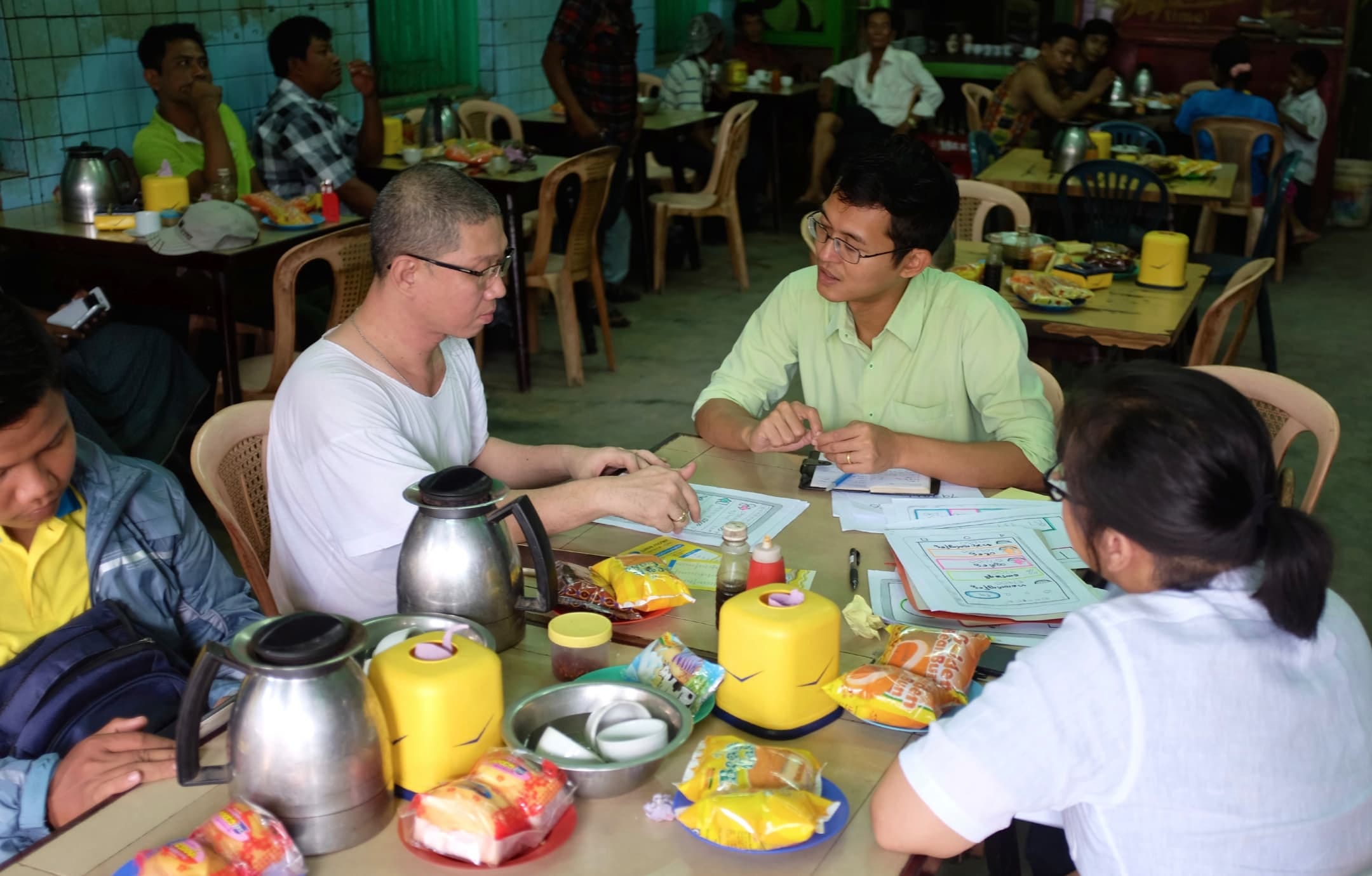 Four people sit at a table featuring packaged snacks, bottles, coffee pots, and letter-size papers, in an informal restaurant. One man, wearing a light-green shirt, is speaking with another man, in a white t-shirt, who is pointing to a paper in front of him. Two other people are in the foreground, one looking down and one with her back turned.