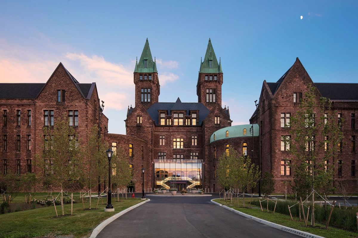 Dusk view of the entry drive leading toward a Romanesque building that has had a glass pavilion inserted at its entrance 