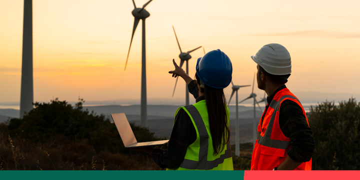 Two energy workers, a woman in a green vest and a man in an orange vest, pointing at wind turbines with their backs to the c.