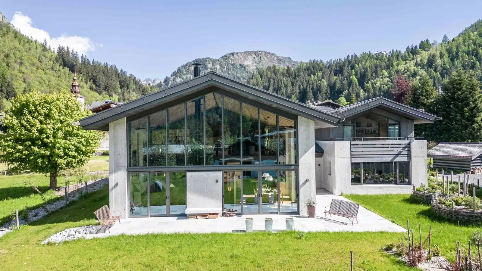 Chalet contemporain avec façade vitrée et vue sur les montagnes, entouré de verdure dans un cadre alpin paisible.
