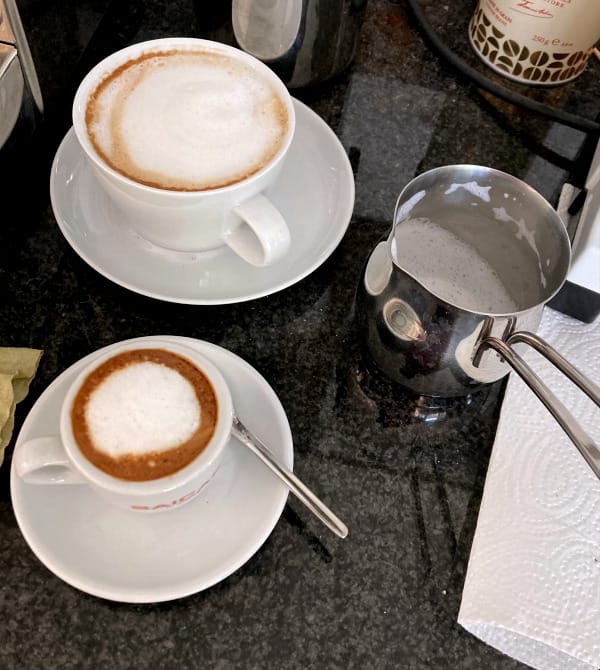 Auf einer schwaren Mamorplatte steht auf UNtertassen eine Espressotasse mit Kaffe und MIlchschaum, dahiner eine Capoccinotasse mit Milchschaum. Daneben ein Edelstahlkännchen mit Henkel und Milchresten