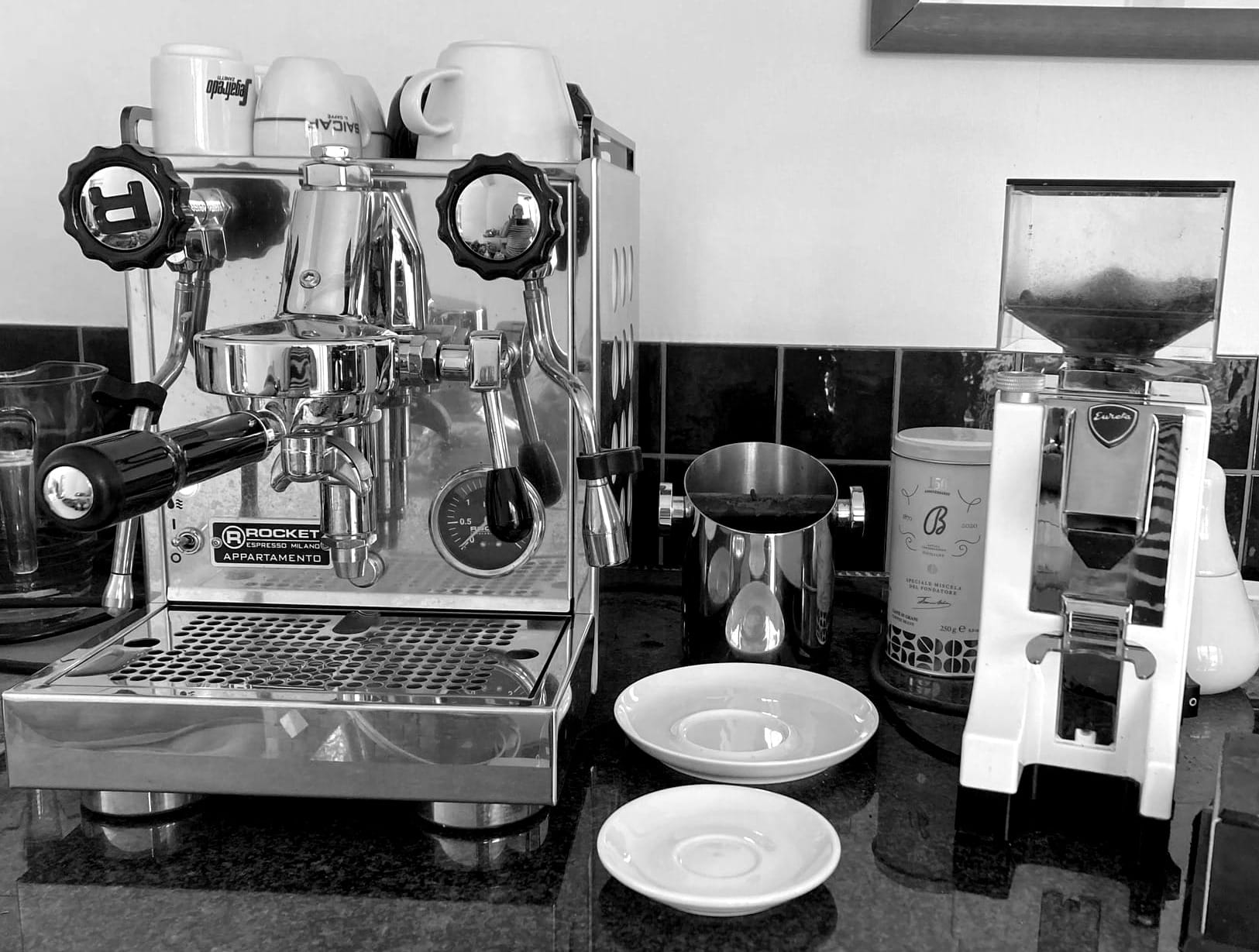 Auf einer schwarzen Mamorarbeitsplatte steht eine Siebträger-Espressomaschine mit Espresso- und Capoccinotassen oben auf dem Deckel. Daneben steht eine große und eine kleine Untertasse. Weiter rechts steht noch eine elektrische Kaffemühle