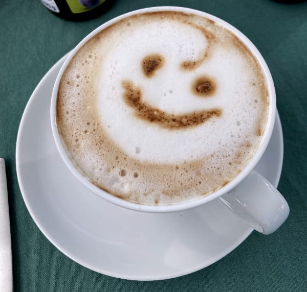 Auf grüner Tischdecke stehend eine weiße Capoccinotasse mit einem lächelnden Gesicht im Milchschaum