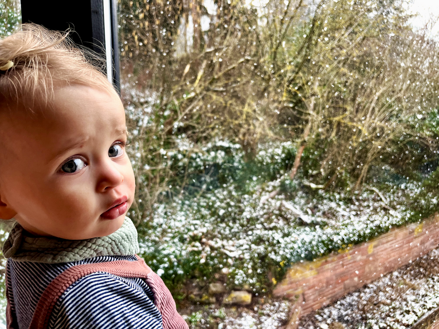 A baby looking skeptically at the camera while it snows outside