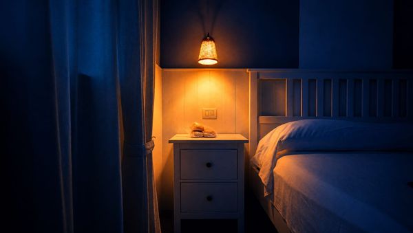 A bedside lamp lighting a nightstand beside a bed at night