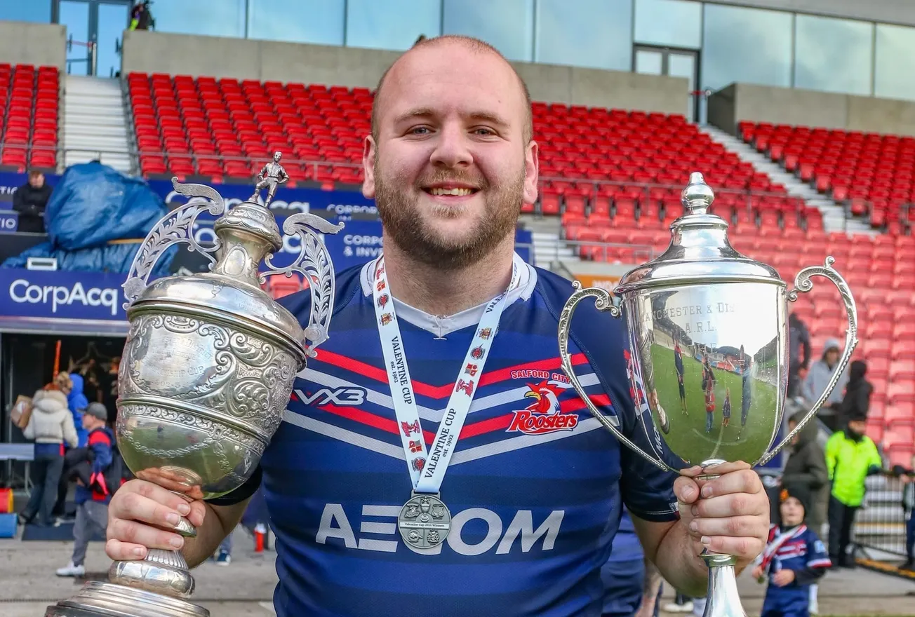 A player wearing a Salford Roosters kit holds two trophies up.