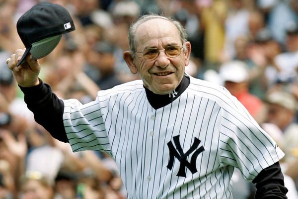 An old Yogi Berra wears a white pinstriped New York Yankees home jersey and waves his cap to the crowd.