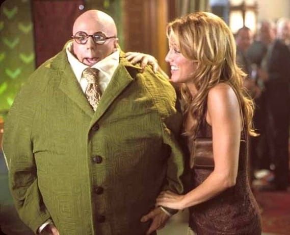 A bald man wears a large, elliptical green suit. He's wearing glasses, and his head is slightly retracting into the suit to emulate a turtle in its shell.
