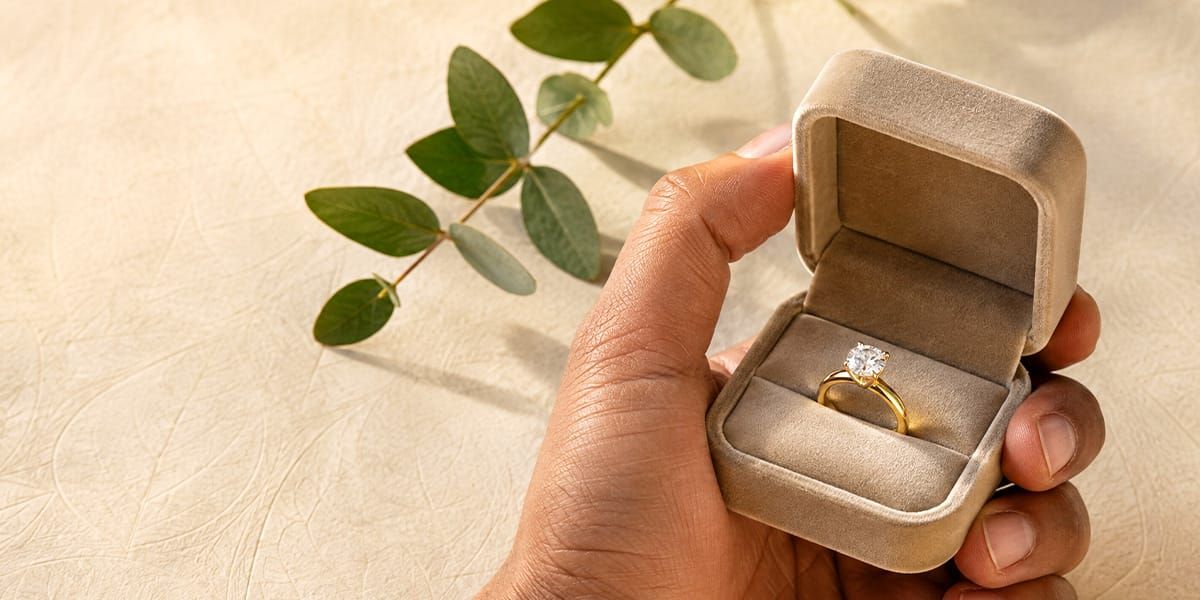 Man's hand presenting a solid gold engagement ring in an open ring box, styled with dried leaves on a warm neutral background