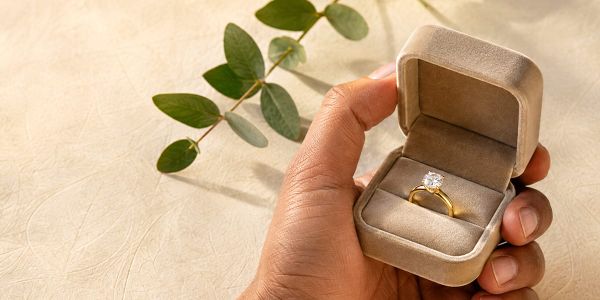 Man's hand presenting a solid gold engagement ring in an open ring box, styled with dried leaves on a warm neutral background