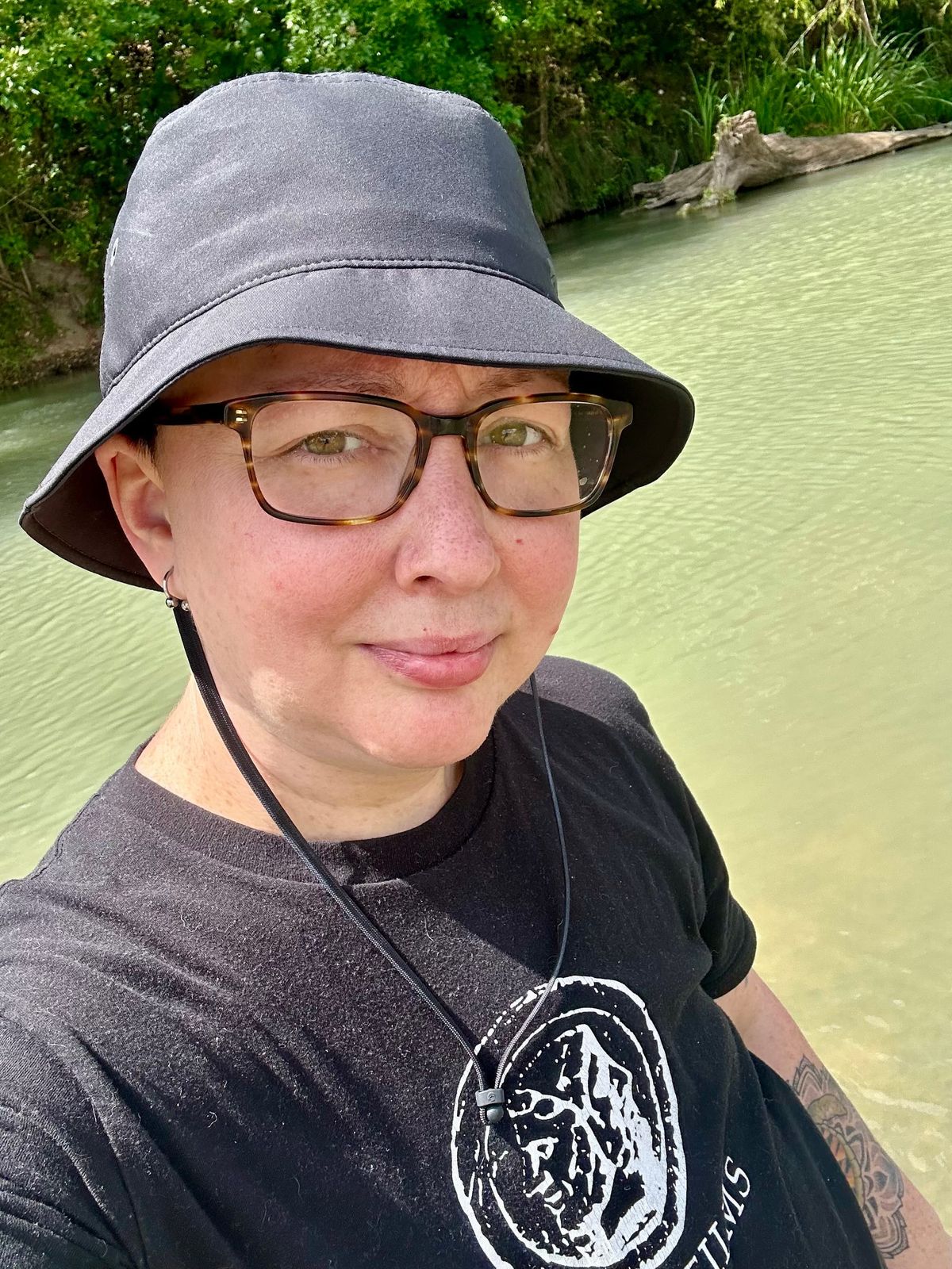 photo of me standing in front of the San Marcos River wearing a black Janus Films t-shirt and black bucket hat.