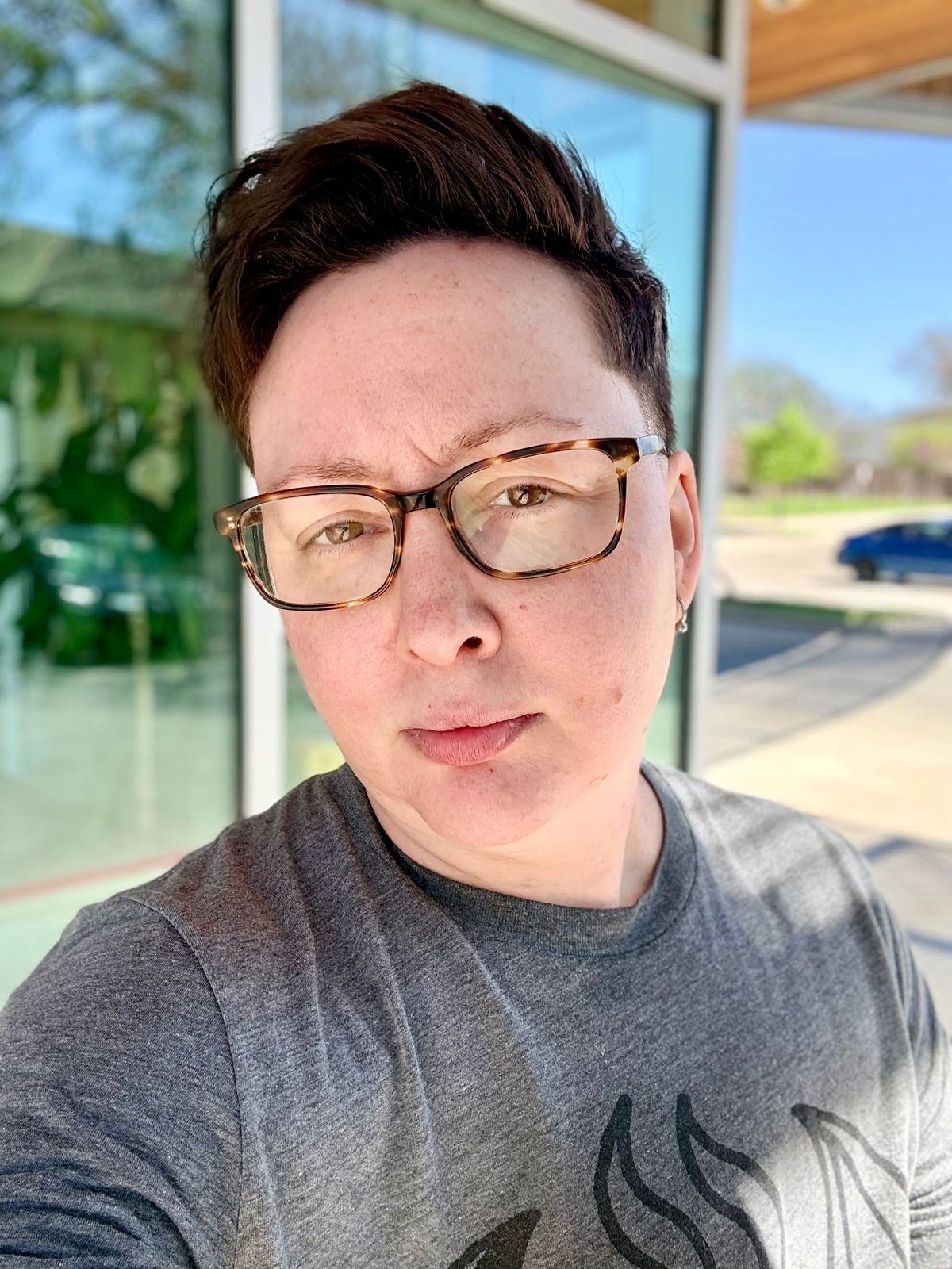 Outdoor selfie photo of A. Max Traphagan, a person with short brown hair wearing tortoiseshell glasses, standing in front of a building with large windows in bright sunlight.
