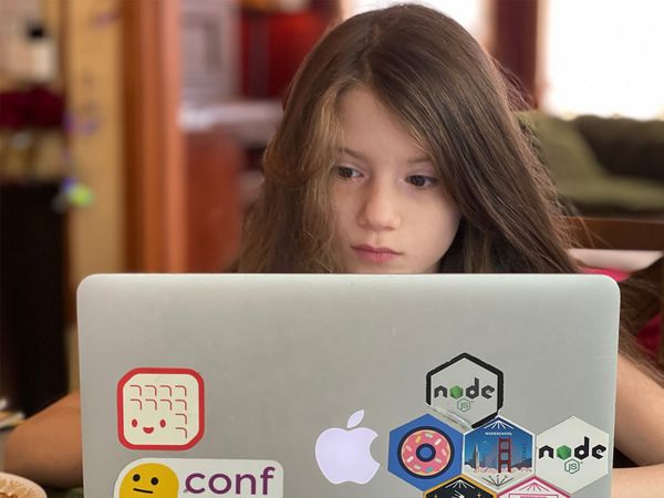 A young girl is working intently at her laptop, which is decorated with Node.js stickers.