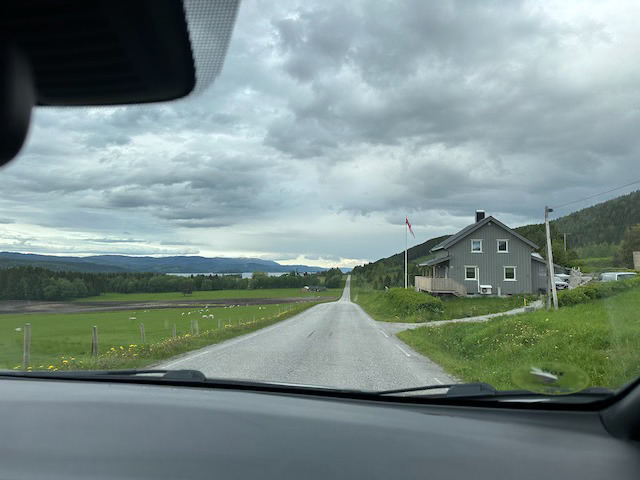 Utsikt gjennom bilrute til landevei som strekker seg rett fram i horisonten, et grått hus langs veien, grønt gress og fjell og vann i det fjerne, med grå skyer over.