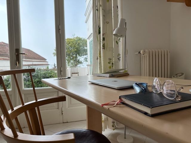 Skrivebord, stol og vindu. Laptop og notatbok med briller på bordplate.
