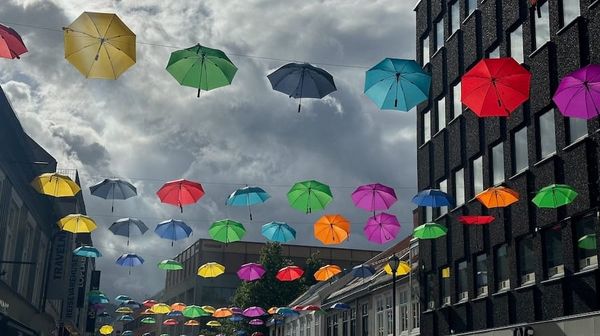 Paraplyer i mange farger henger på snor mellom husene i en bygate med grå uværsskyer i bakgrunnen.