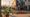 Golden-hour LA side street with murals, café tables, locals chatting, and palm shadows stretching across sunlit sidewalks.