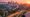 Golden-hour view of Los Angeles skyline from hilltop, with glowing palms, amber light, and snowcapped mountains under a pink-violet sky.