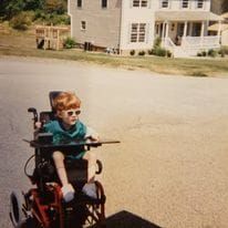 An image of me as a young child in a red wheelchair. I have bright red hair and I'm wearing sunglasses.