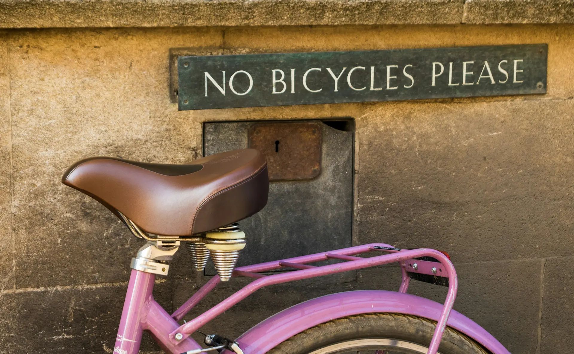 Photo of a sign on a building that says “No Bicycles Please” with a pink bicycle right under the sign.