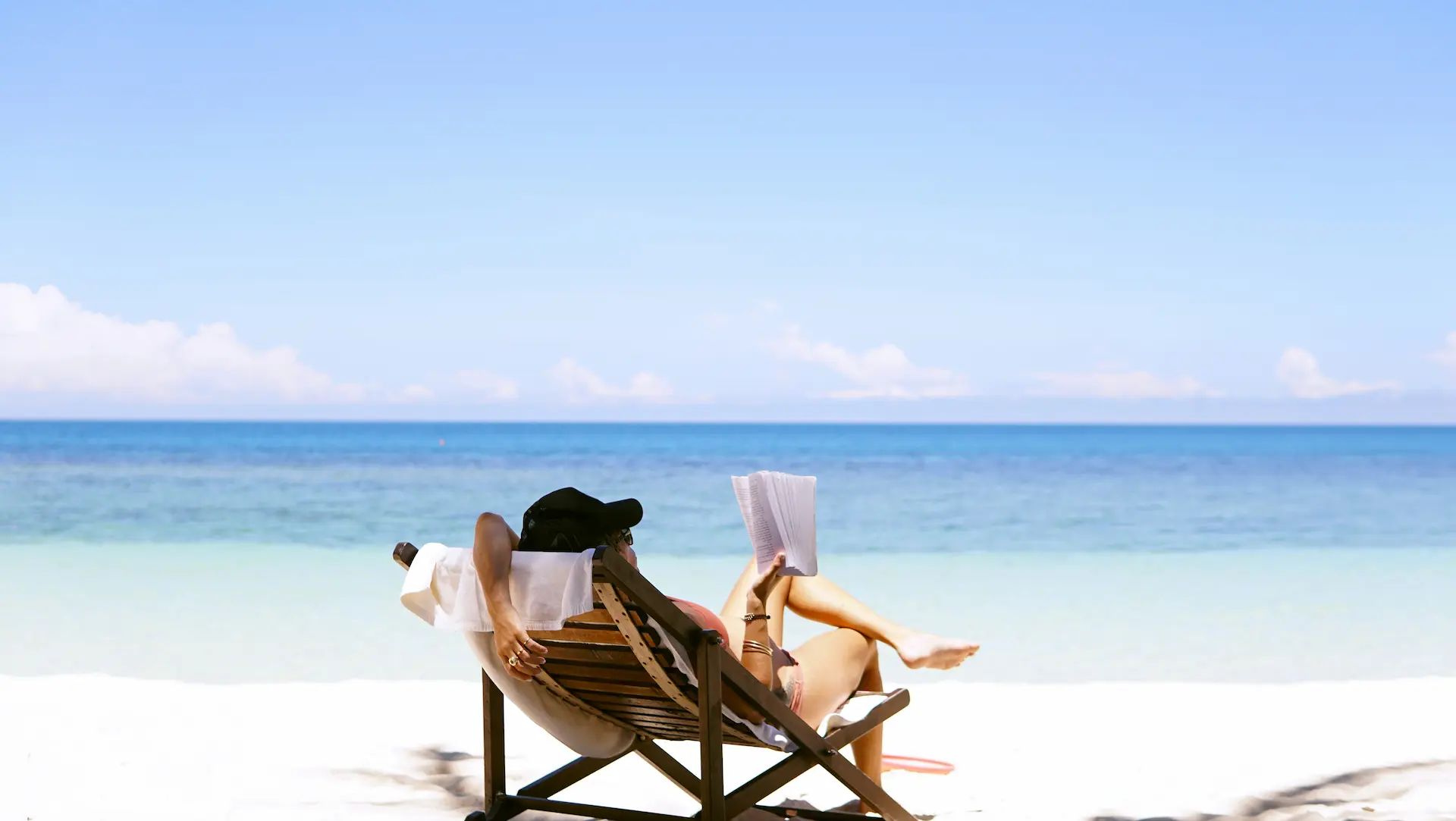 Woman reading a book on a wooden sun lounger at the sea on a white sand beach.