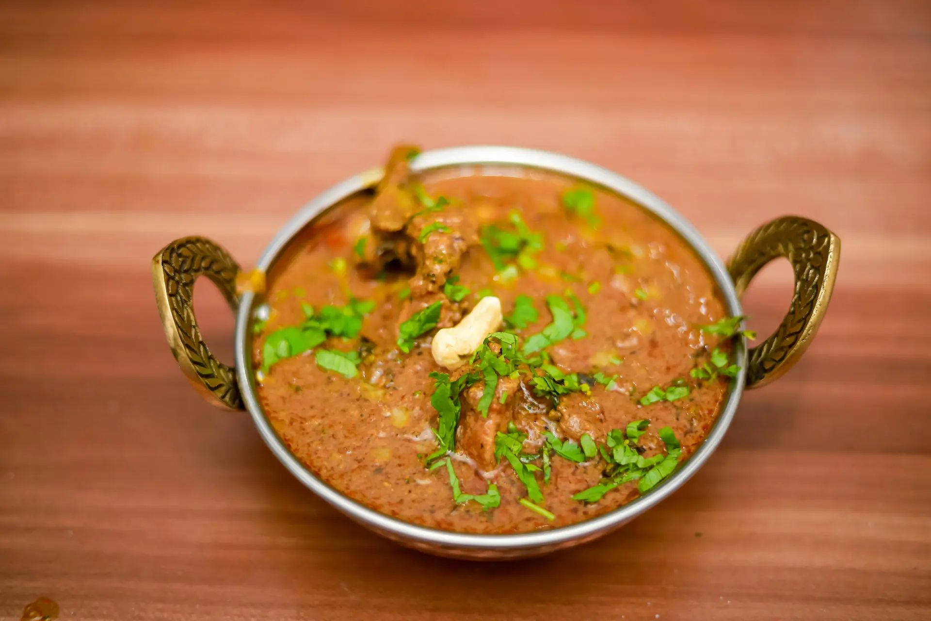 Photo of a curry in a beautifully decorated brass bowl. Probably a lamb rogan josh with cilantro and a single cashew nut.