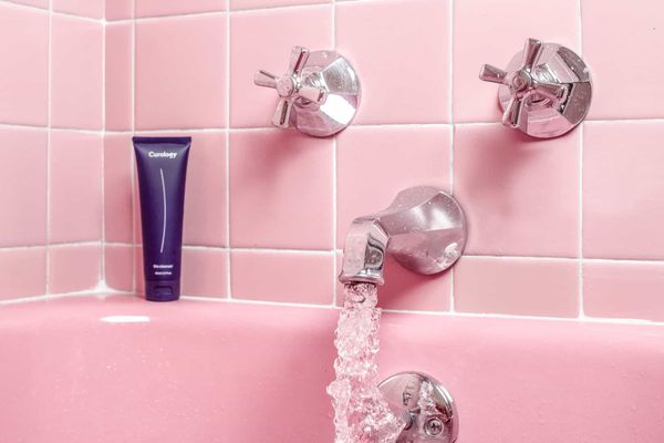 A close up photo of a tap and two knobs of a very pink bathtub in a very pink tiled bathroom. Water flows out of the tap.