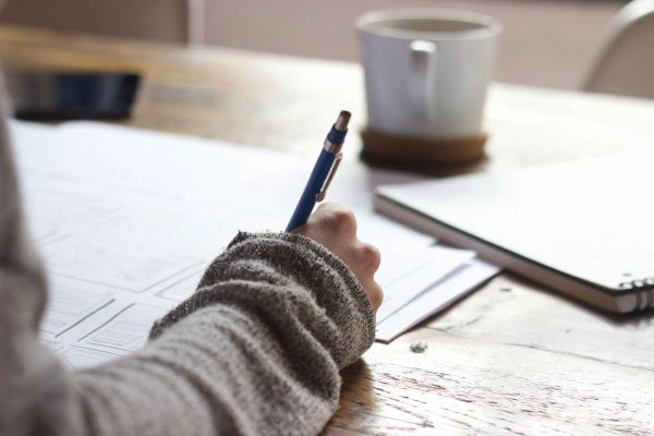 Person taking notes on paper while sitting at a wooden table. A blurry mug and a notebook are in the background