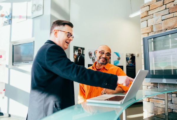 Two men looking at a laptop in a brightly lit modern art gallery. One of them is explaining something to the other.